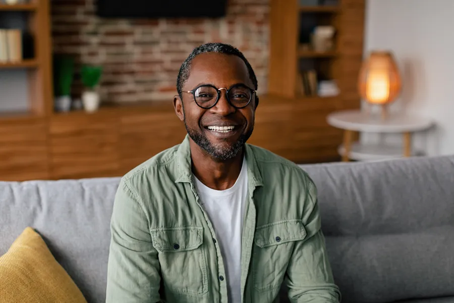 Andropause-Doctor A middle aged black man in a green button up shirt sits on his couch smiling, happy with his treatment for andropause and low testosterone from Tanya Zboril, NP of TBT Medical in Kitchener-Waterloo.