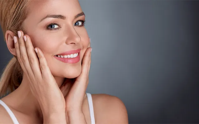 Facial-Clinic A woman with clear, radiant skin smiling at the camera with both hands on her cheeks. Representing Facials offered by Tanya Zboril, NP of TBT Medical in Kitchener-Waterloo.
