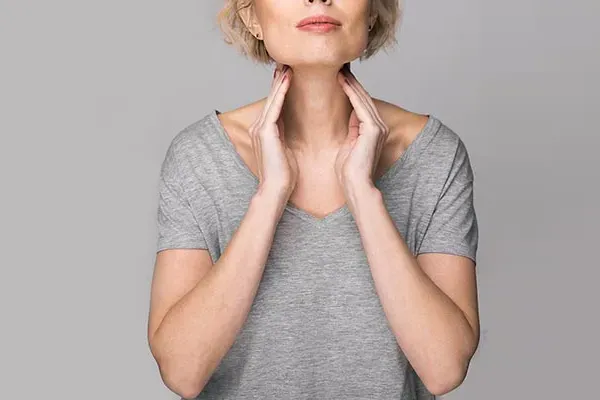 Hashimotos-Thyroiditis-Doctor An older blonde woman in a gray t-shirt holds her hands to her neck checking for signs of thyroid problems. Get help for Hashimoto's Thyroiditis from Tanya Zboril, NP of TBT Medical in Kitchener-Waterloo.