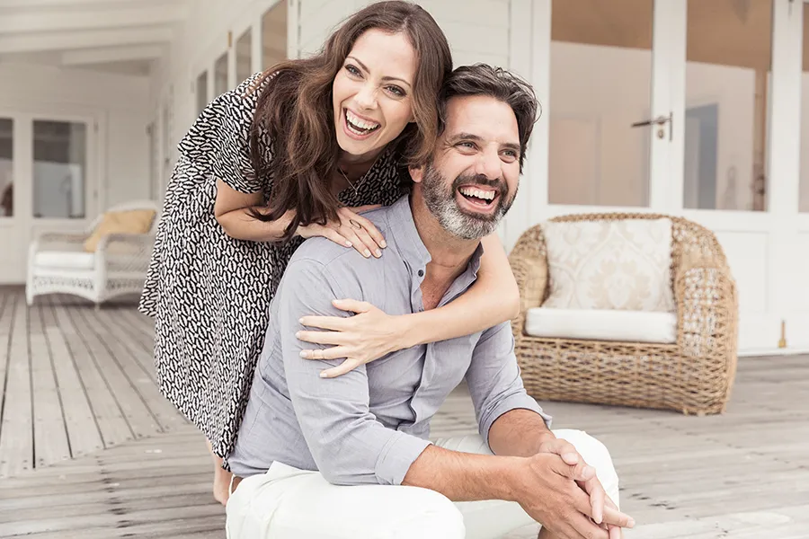 Health-and-Wellness-Services-Doctor A middle-aged couple on a wooden porch with wicker furniture; the woman is playfully hugging the man from behind. They are benefiting from the health and wellness services from Tanya Zboril, NP of TBT Medical in Kitchener-Waterloo.