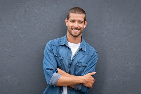 Hormones-for-Men-Doctor A man in a denim shirt stands smiling against a gray-blue wall, pleased with his testosterone hormone therapy from Tanya Zboril, NP of TBT Medical in Kitchener-Waterloo.