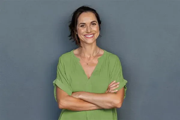 Hormones-for-Women-Doctor A woman in a green shirt stands smiling against a gray-blue wall, pleased with her hormone therapy from Tanya Zboril, NP of TBT Medical in Kitchener-Waterloo.