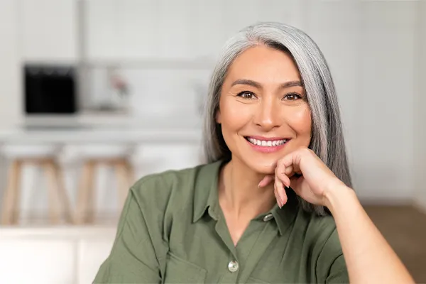 Menopause-Clinic An asian woman with long gray hair and a green shirt sitting and smiling on her couch, happy with her menopause care from [NAME} in Kitchener-Waterloo.
