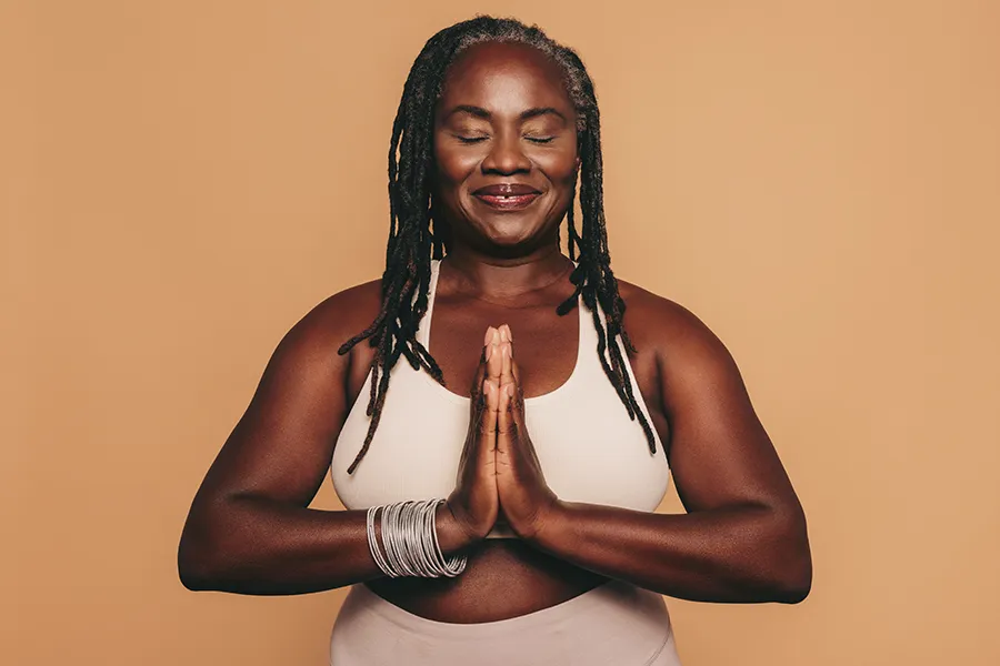 Obesity-Clinic An older African American woman confidently posing against a warm background smiling with hands in a prayer pose. Tanya Zboril, NP of TBT Medical treats obesity using functional medicine in Kitchener-Waterloo.