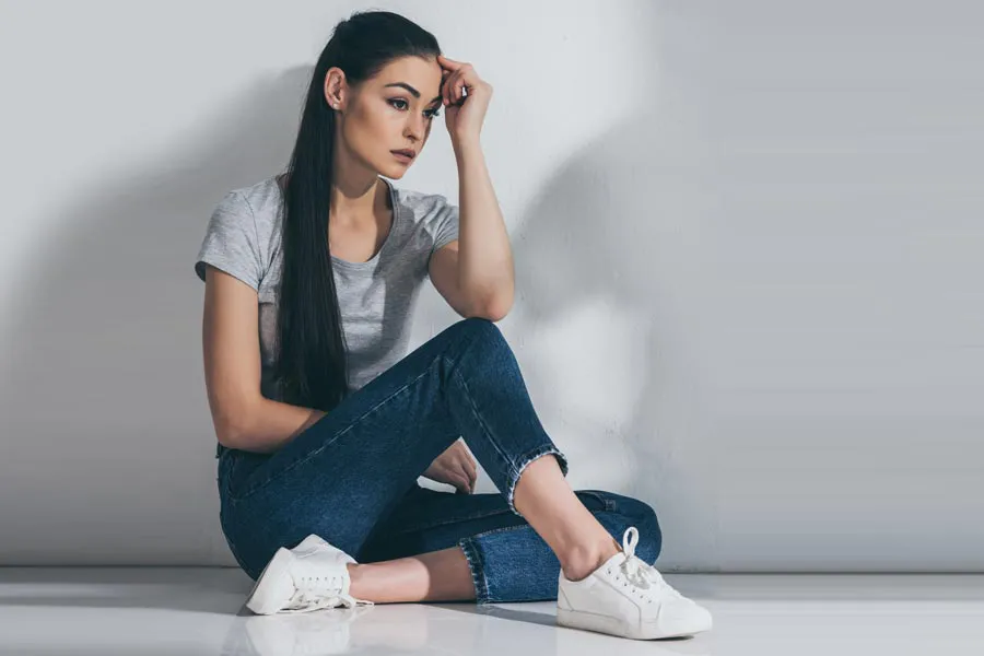 PCOS-Doctor A young brunette woman in a gray t-shirt and jeans sits against a gray wall with her head resting on her hand and a distressed expression. Get help for Polycystic Ovarian Syndrome (PCOS) from Tanya Zboril, NP of TBT Medical in Kitchener-Waterloo.
