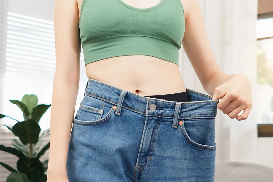 Zepbound-Weight-Loss-Clinic A close-up of a young woman in a green crop top, holding out her oversized blue jeans to show significant weight loss. Zepbound Weight Loss Injections offered by Tanya Zboril, NP of TBT Medical in Kitchener-Waterloo.
